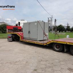 SecureTrans - Szara szafa elektryczna zabezpieczona łańcuchami na żółto-czerwonej niskopodwoziowej naczepie ciągniętej przez białą ciężarówkę, logo firmy transportowej w rogu