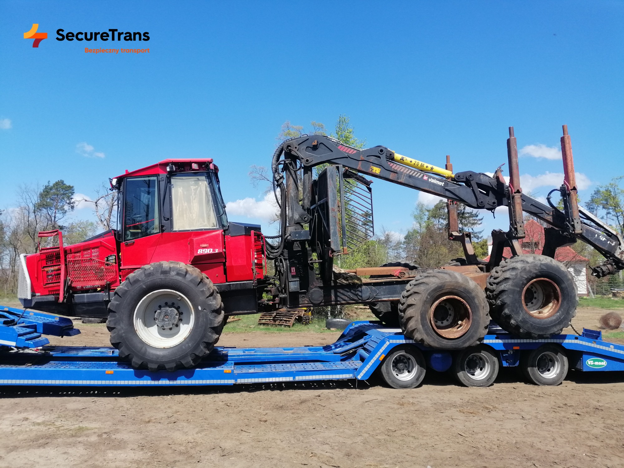 Czerwony harwester Valmet 890.3 z chwytakiem, transportowany na niskopodwoziowej, niebieskiej lawecie VS-mont, na tle polnej drogi i błękitnego nieba.