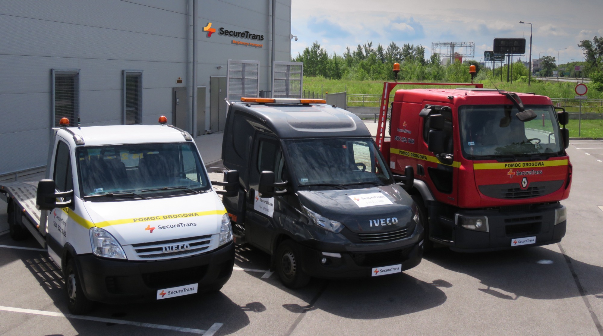 Trzy pojazdy Iveco w kolorach białym, czarnym i czerwonym z napisem 'Pomoc Drogowa' i logo SecureTrans, stojące na parkingu przed budynkiem firmy z podobnym logo.