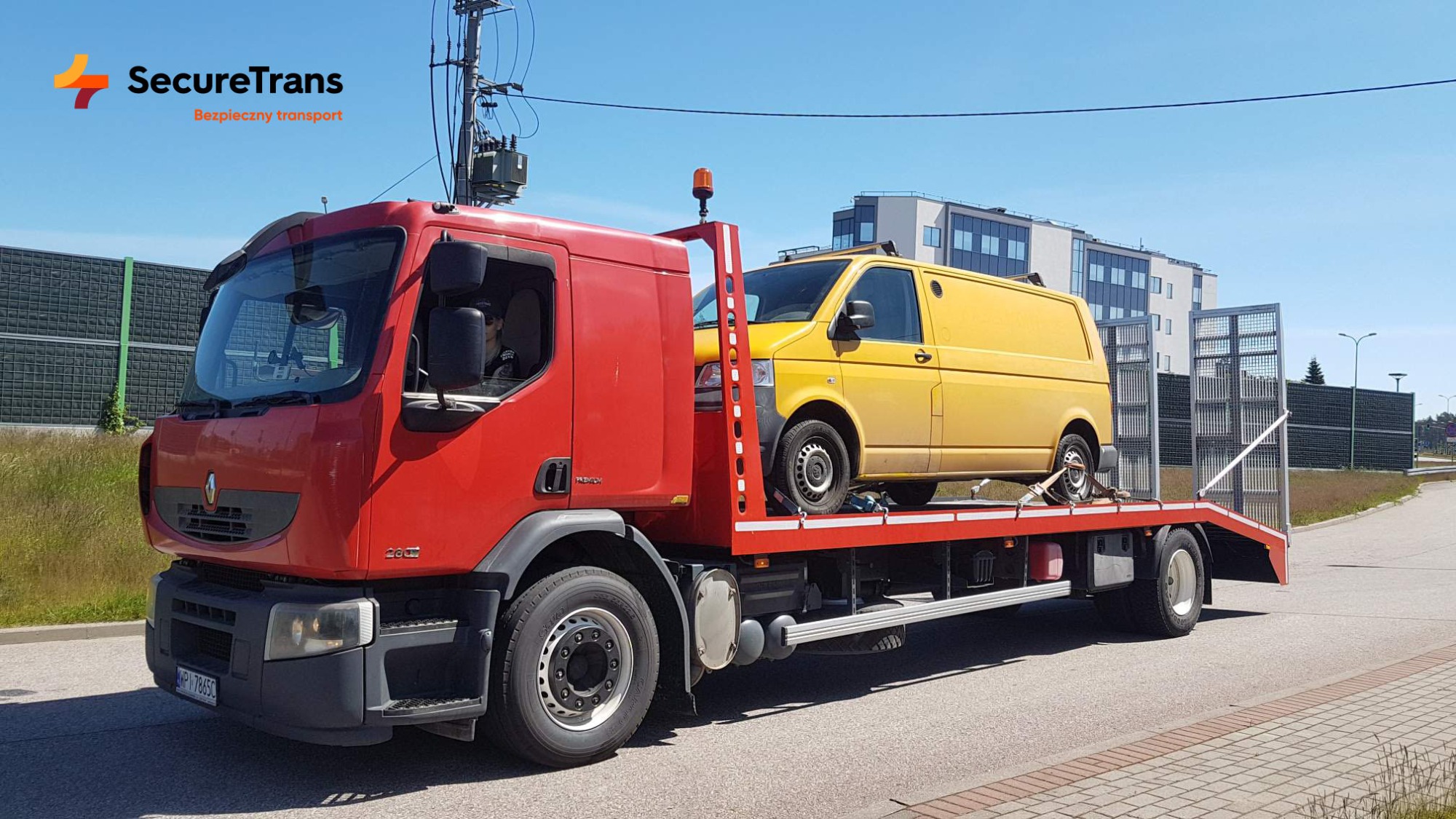 Czerwona laweta Renault z żółtym busem Volkswagen na platformie, zabezpieczonym pasami transportowymi, w tle budynek biurowy i bariera akustyczna.