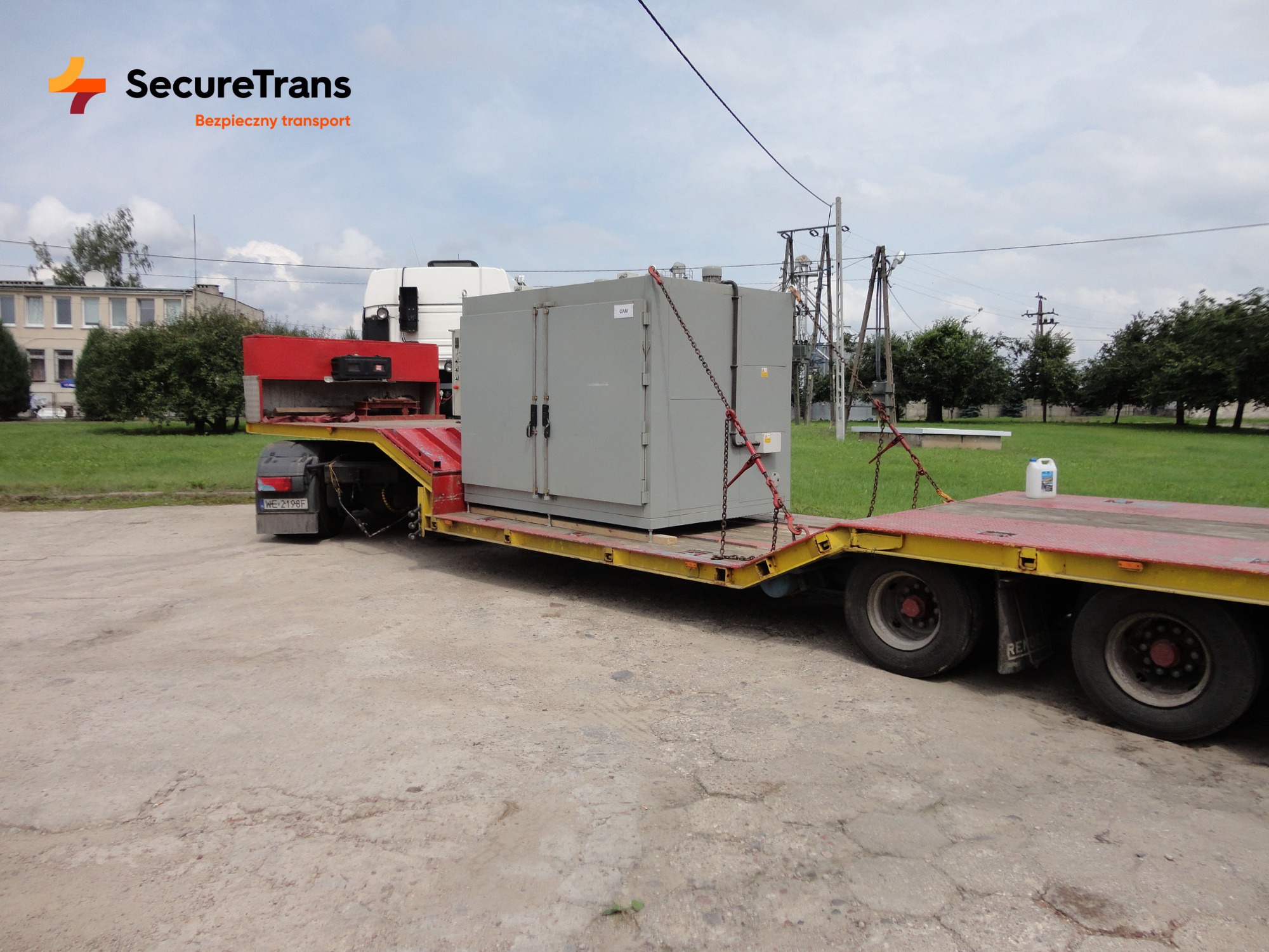 Szara szafa elektryczna zabezpieczona łańcuchami na żółto-czerwonej niskopodwoziowej naczepie ciągniętej przez białą ciężarówkę, logo firmy transportowej w rogu