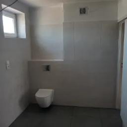 Wykończona łazienka z szarymi płytkami na ścianach i podłodze, wisząca toaleta i widoczna kratka wentylacyjna pod sufitem.