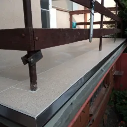 Wyłożony jasnymi płytkami taras z brązową balustradą i metalowymi słupkami, widok z dołu, z fragmentem ceglanej ściany w tle.