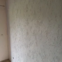 Ściana wewnętrzna z dekoracyjną fakturą w odcieniach szarości, widoczna z niskiego kąta, z fragmentem podłogi z paneli i białym narożnikiem ściany.