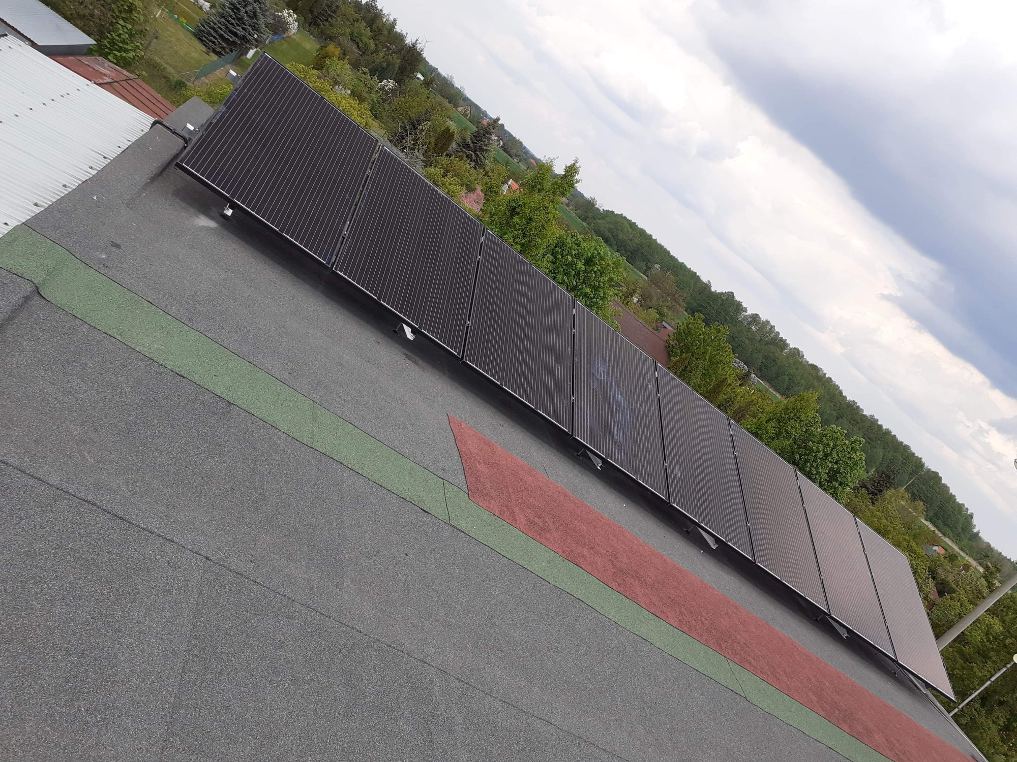 Rząd paneli fotowoltaicznych na płaskim dachu pokrytym papą z zielonymi i czerwonymi pasami, widok z góry na tle zieleni i pochmurnego nieba.
