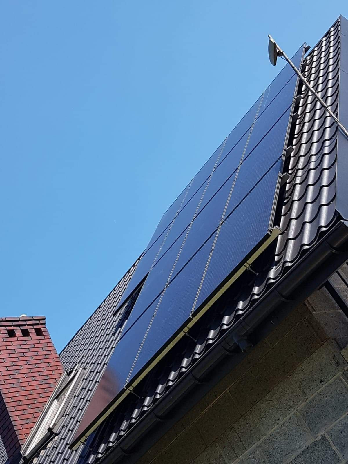 Panele słoneczne na dachu z czarnej dachówki, widok z dołu, na tle błękitnego nieba, widoczny komin z czerwonej cegły.