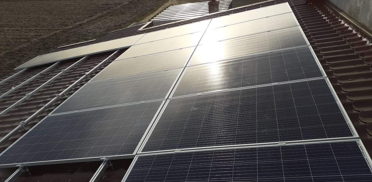 Montaż paneli słonecznych na dachu z brązową dachówką, widoczne metalowe konstrukcje mocujące i częściowo zamontowane panele, odbicie słońca na powierzchni paneli.