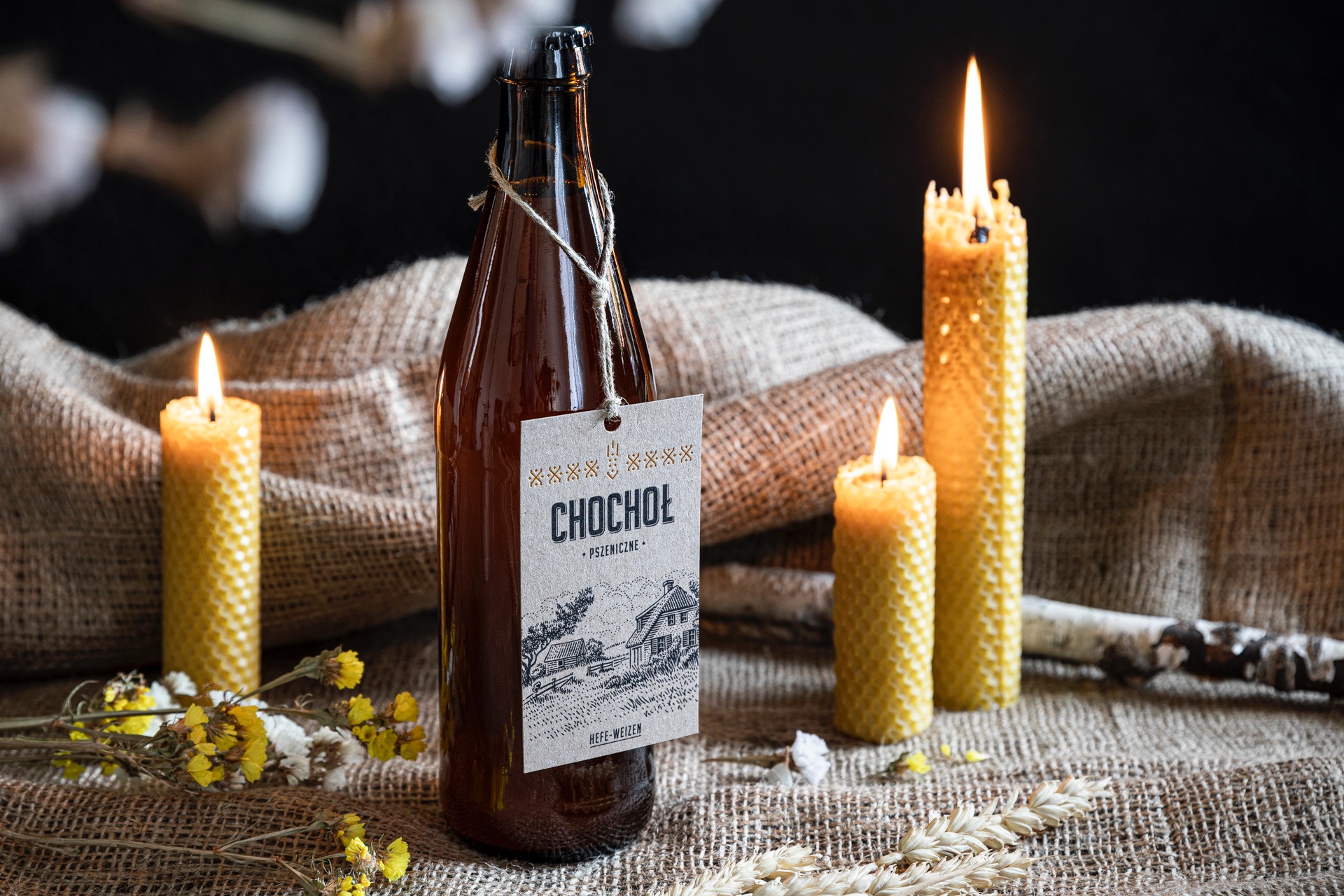 Butelka piwa rzemieślniczego 'Chochoł' z etykietą przedstawiającą wiejski krajobraz, otoczona płonącymi świecami z wosku pszczelego i suszonymi kwiatami na tle lnianej tkaniny.
