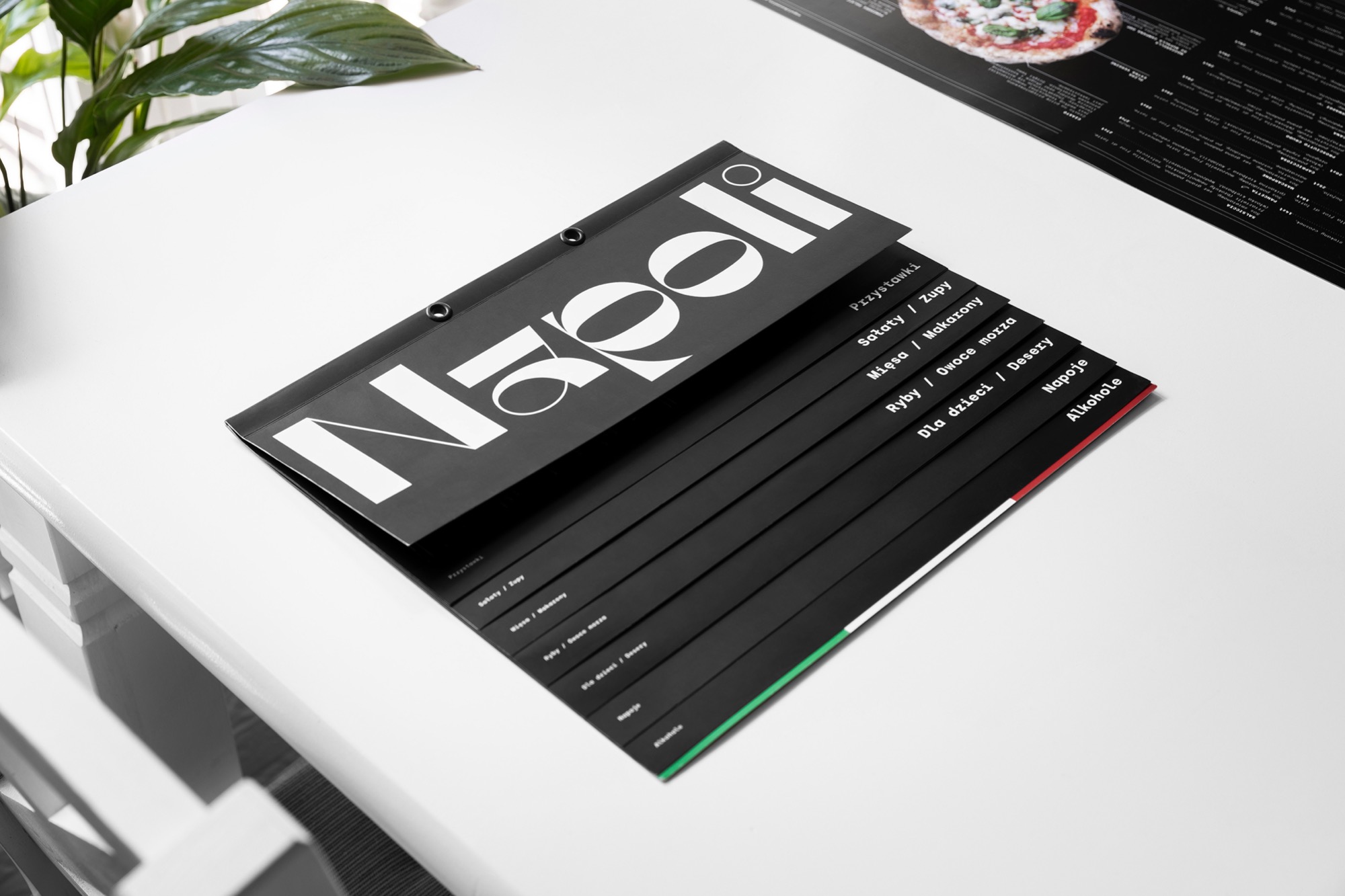Elegancko zaprojektowane menu restauracji Napoli, z czarnymi kartami i białym, nowoczesnym fontem, leżące na białym stole obok zielonej rośliny doniczkowej.