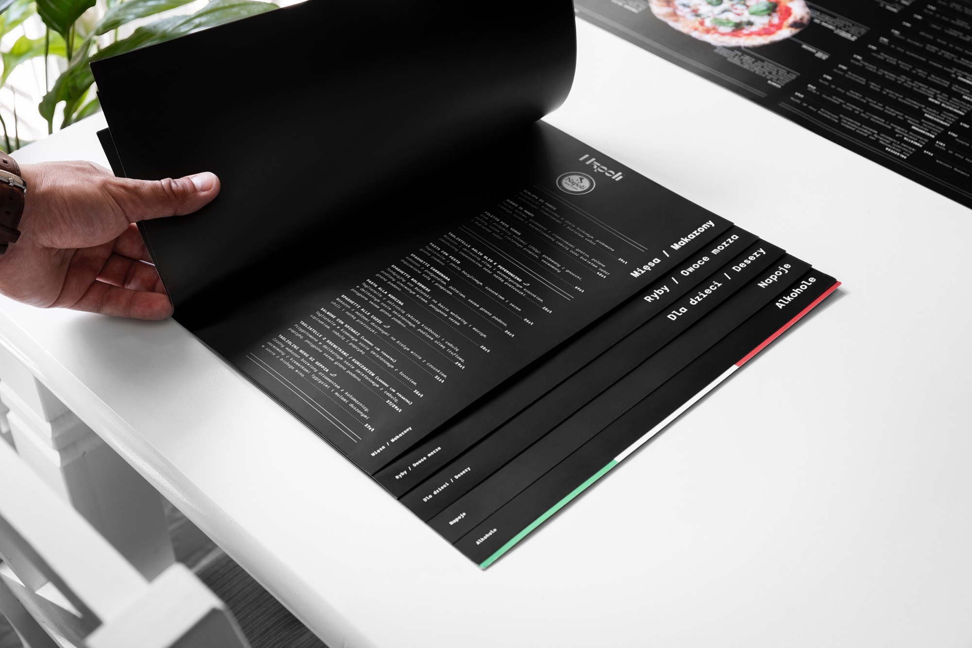 Otwarta, czarno-biała karta menu restauracji włoskiej z wyraźnie widocznymi kategoriami dań: mięsa/makarony, ryby/owoce morza, dla dzieci/desery, napoje, alkohole. Na stronie z daniami szczegółowy...