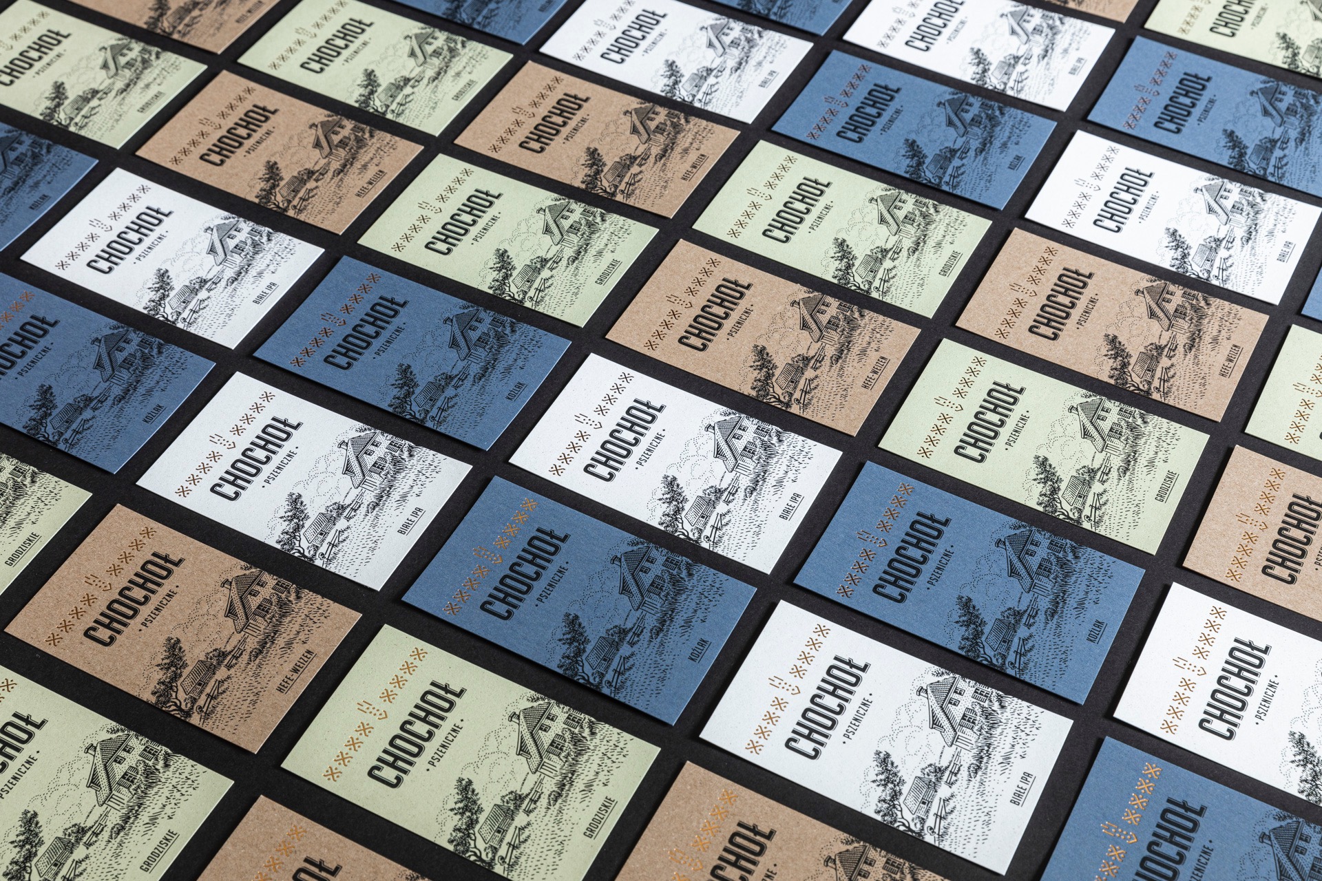 Różnokolorowe wizytówki z nazwą browaru 'Chochoł' i rysunkiem wiejskiej chaty, ułożone pod kątem na czarnym tle. Każda wizytówka ma inny odcień: biały, niebieski, zielony, brązowy.