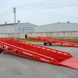 Dwie czerwone rampy przeładunkowe AUSBAU na betonowym placu, z widocznym numerem telefonu i adresem strony internetowej, na tle szarego budynku magazynowego i betonowego ogrodzenia.