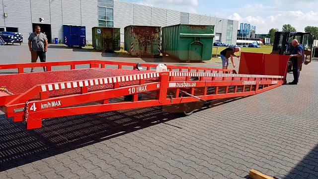 Czerwony, mobilny najazd rampowy z kratownicą, ustawiony na placu z kostki brukowej, przygotowywany do użycia przez dwóch mężczyzn, z wózkiem widłowym w tle, obok widoczne kontenery.