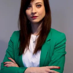 Portret kobiety w zielonej marynarce, z rękami skrzyżowanymi na piersiach i srebrnym naszyjnikiem z sercem, patrzącej w górę.