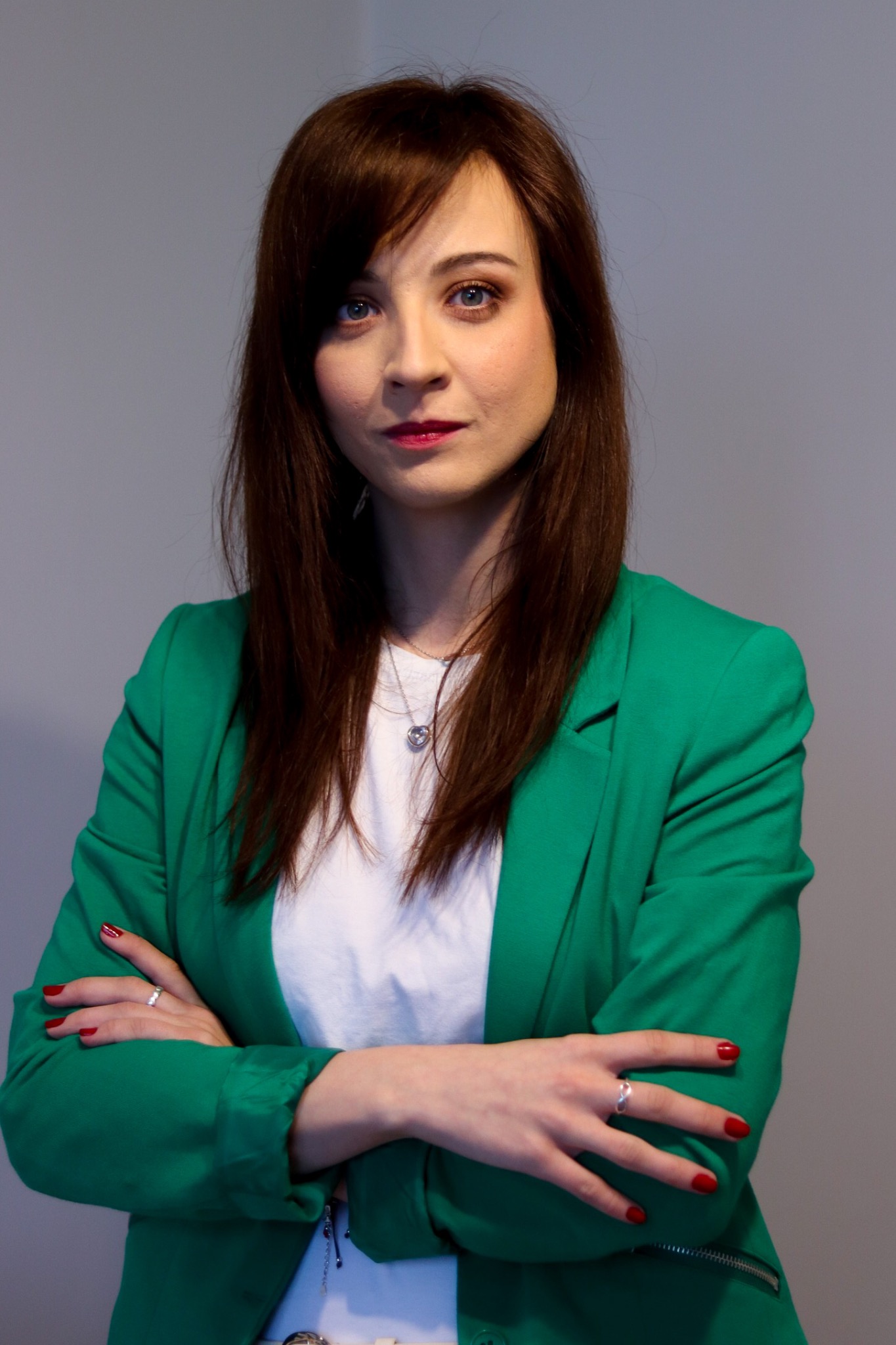 Portret kobiety w zielonej marynarce, z rękami skrzyżowanymi na piersiach i srebrnym naszyjnikiem z sercem, patrzącej w górę.