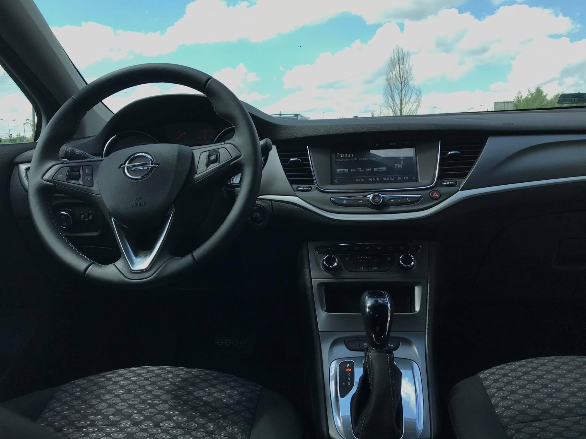Wnętrze samochodu marki Opel z automatyczną skrzynią biegów, widok na deskę rozdzielczą, kierownicę z logo, radio ustawione na stację z Poznania i widok na niebo przez przednią szybę.