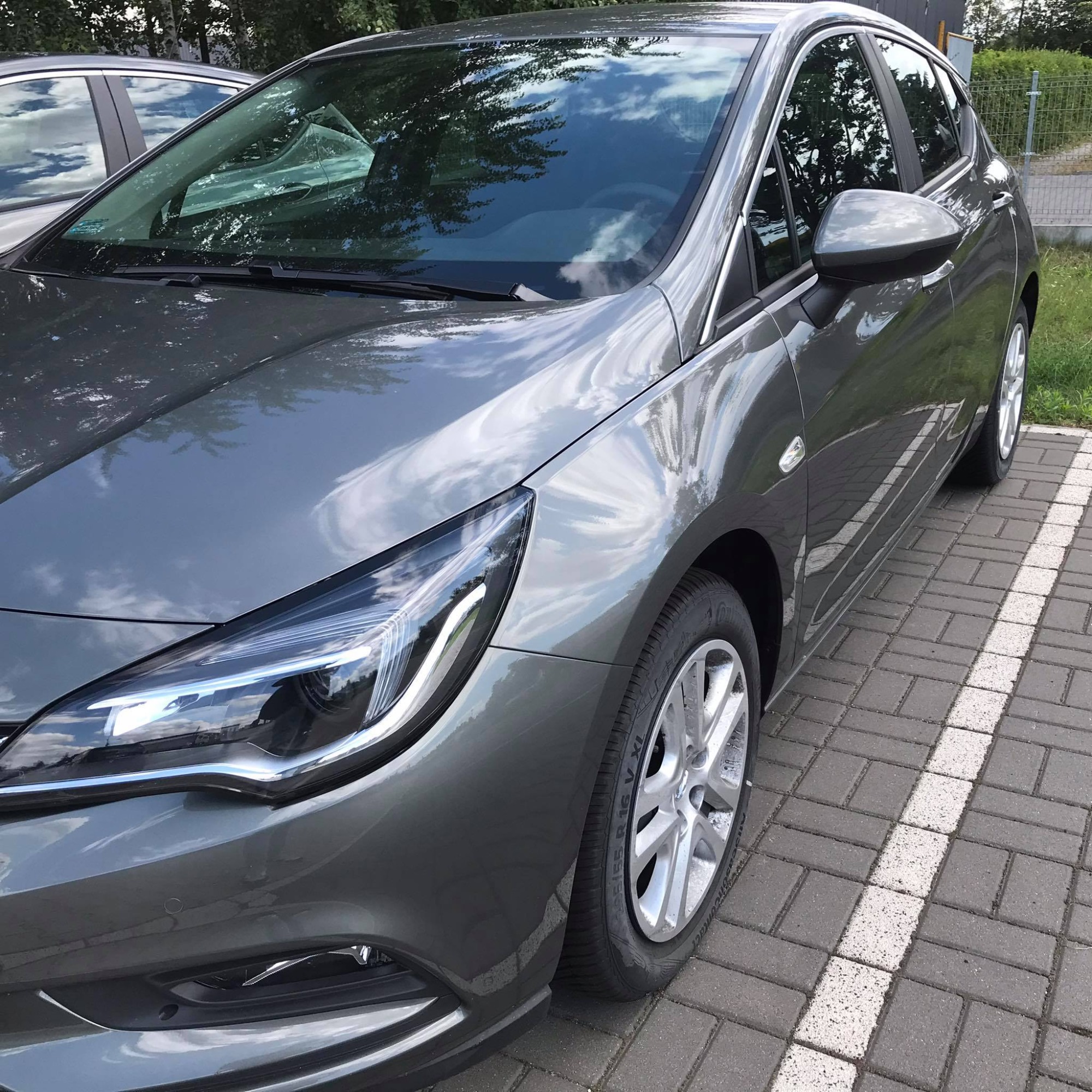 Szary samochód osobowy zaparkowany na parkingu z kostki brukowej, widok z boku na przedni reflektor i koło, z odbiciami chmur na karoserii.