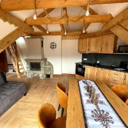 Wnętrze góralskiego domu z drewnianą konstrukcją stropu, kamiennym kominkiem i kuchnią z drewnianymi szafkami, oświetlone wiszącymi żarówkami na linach.