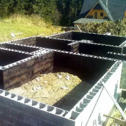 Zalane fundamenty z bloczków betonowych, zabezpieczone izolacją, widok na dom w budowie i zalesione wzgórze w tle.
