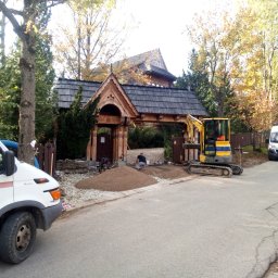 Art Kop - Mini koparka na tle drewnianej bramy, robotnik wyrównujący żwirowe podłoże przed bramą, częściowo widoczny biały samochód dostawczy