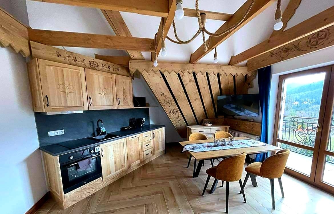 Jasna, drewniana kuchnia z czarnym blatem i wzorzystymi rzeźbieniami na szafkach, widok na stół jadalny i okno z widokiem na góry.