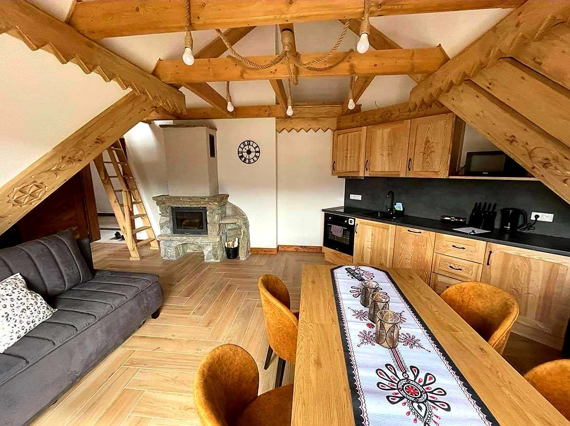 Wnętrze góralskiego domu z drewnianą konstrukcją stropu, kamiennym kominkiem i kuchnią z drewnianymi szafkami, oświetlone wiszącymi żarówkami na linach.