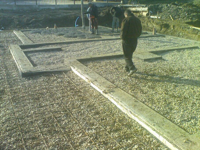 Przygotowanie zbrojenia i szalunku pod wylewkę fundamentów na żwirowym podłożu, widoczni pracownicy w tle.