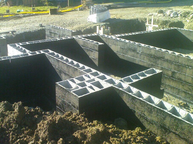 Szare bloczki betonowe tworzące zarys fundamentów budynku, widoczne z ziemi, z żółtymi rurami w tle.