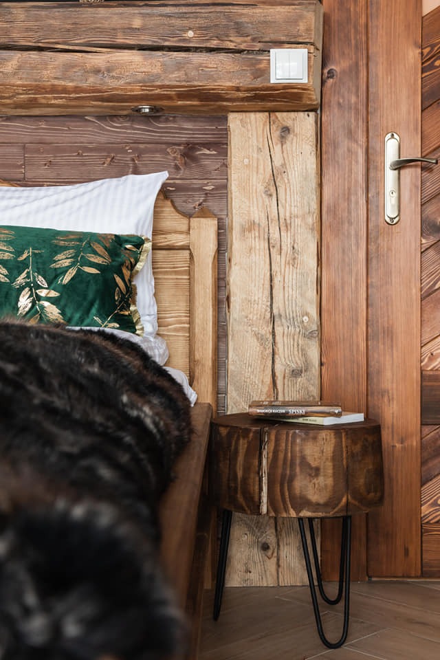 Fragment sypialni w stylu góralskim: drewniane ściany, łóżko z zieloną poduszką i stolik nocny z pnia drzewa na metalowych nóżkach, na którym leżą książki.