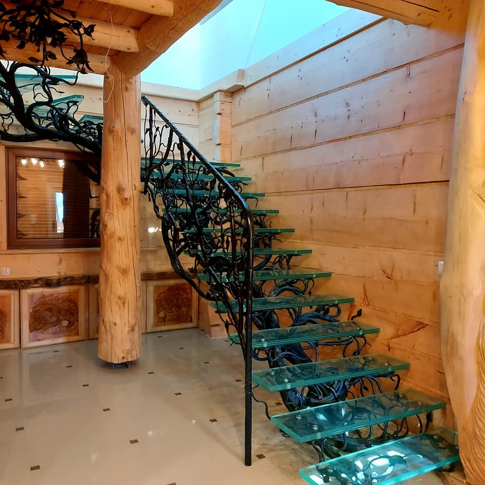 Schody wewnętrzne z przezroczystymi stopniami i metalową balustradą w kształcie gałęzi drzew, w drewnianym domu z widocznym oknem i obrazami na ścianie.