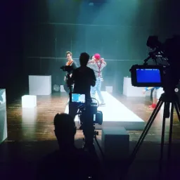 Ekipa filmowa kręci scenę z aktorami na planie: dwie kamery, oświetlenie sceniczne i rekwizyty w postaci białych sześcianów.