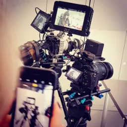 Profesjonalny sprzęt do wideofilmowania: dwie kamery na statywach z monitorami podglądowymi, z których jeden pokazuje scenę z kobietą siedzącą przy biurku, a drugi z perspektywy pierwszej kamery;...