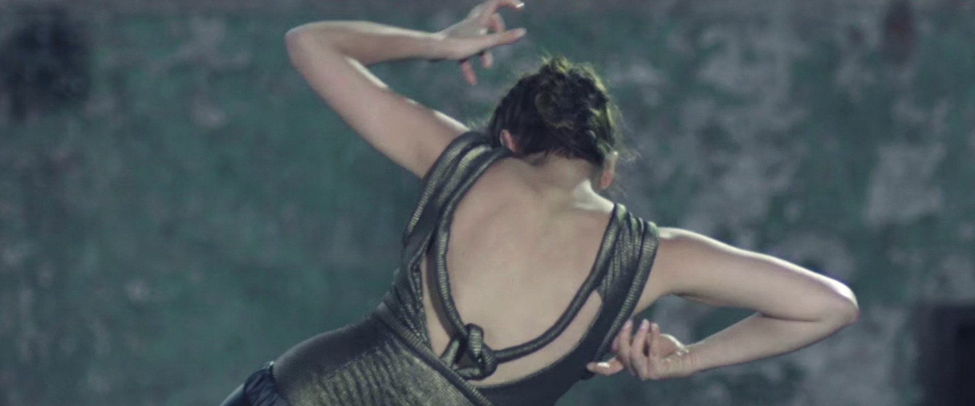 Kobieta w ruchu, widok z tyłu, ubrana w błyszczącą sukienkę z oryginalnym wycięciem na plecach, w trakcie wykonywania układu tanecznego na tle szarej ściany.