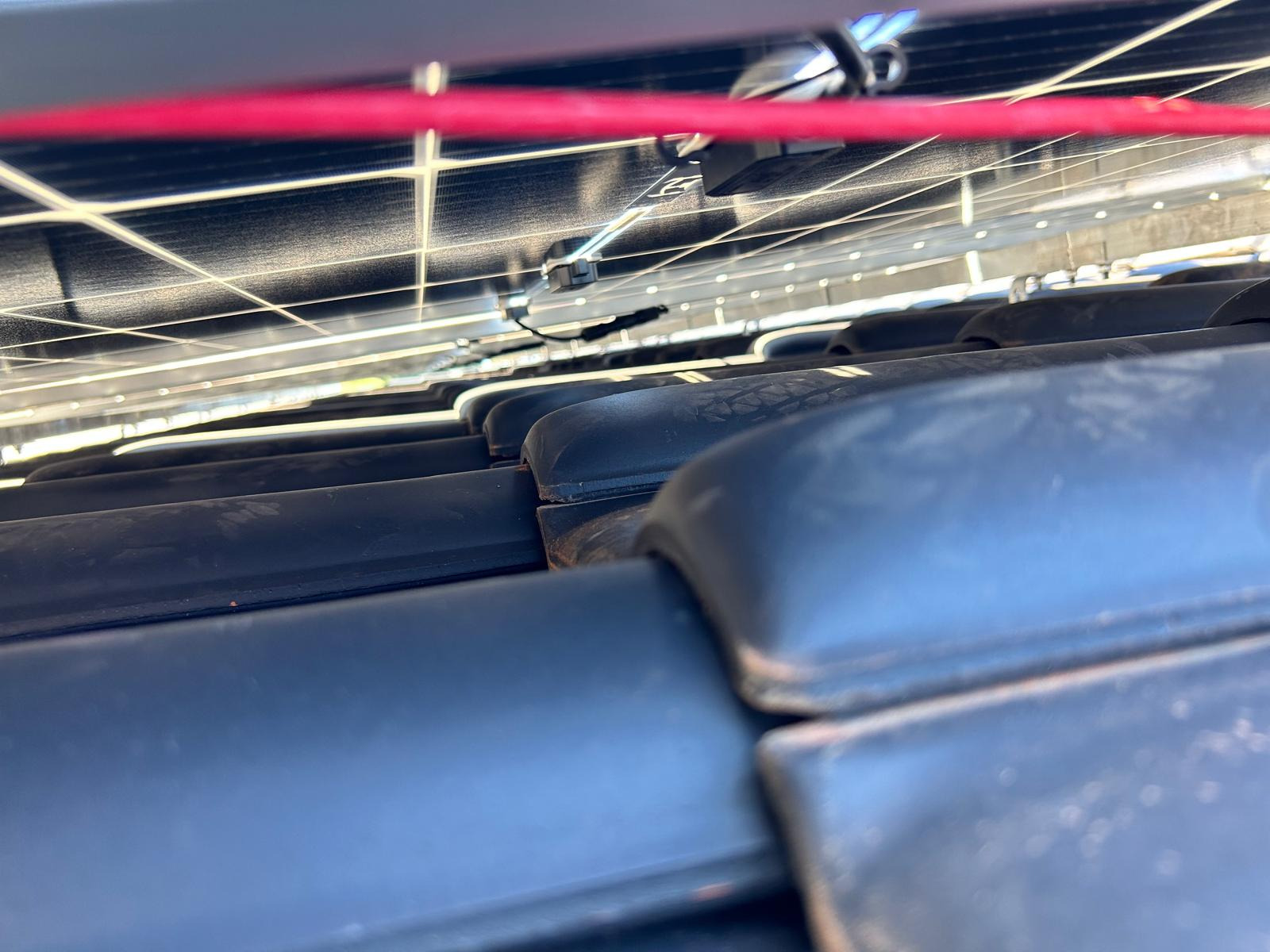 Niski kąt ujęcia paneli fotowoltaicznych zamontowanych na ciemnej dachówce ceramicznej, widoczne kable i elementy montażowe.