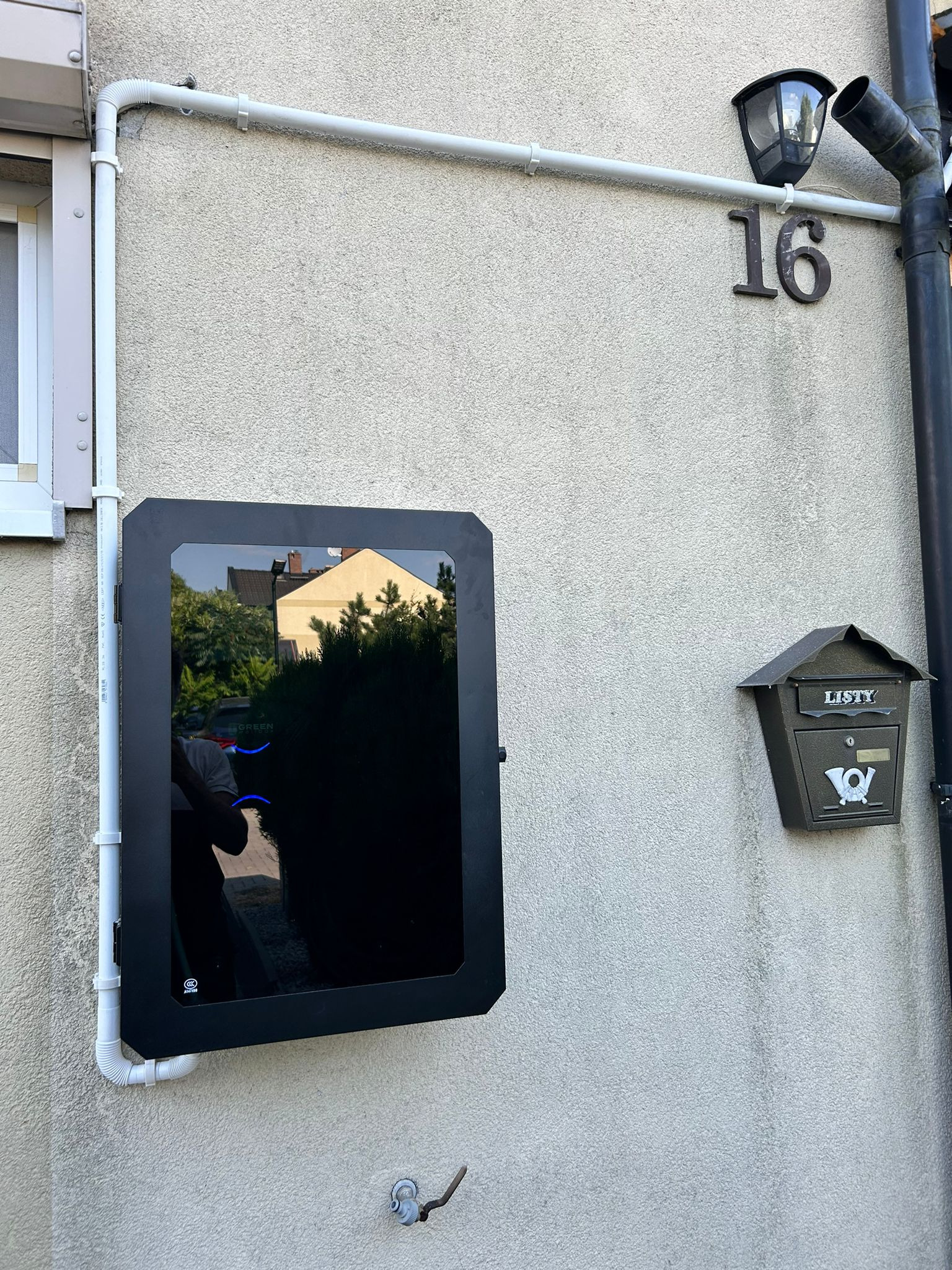 Czarny, obudowany falownik fotowoltaiczny zamontowany na szarej ścianie budynku obok skrzynki na listy i numeru domu 16, z widocznym odbiciem otoczenia w szklanej obudowie.