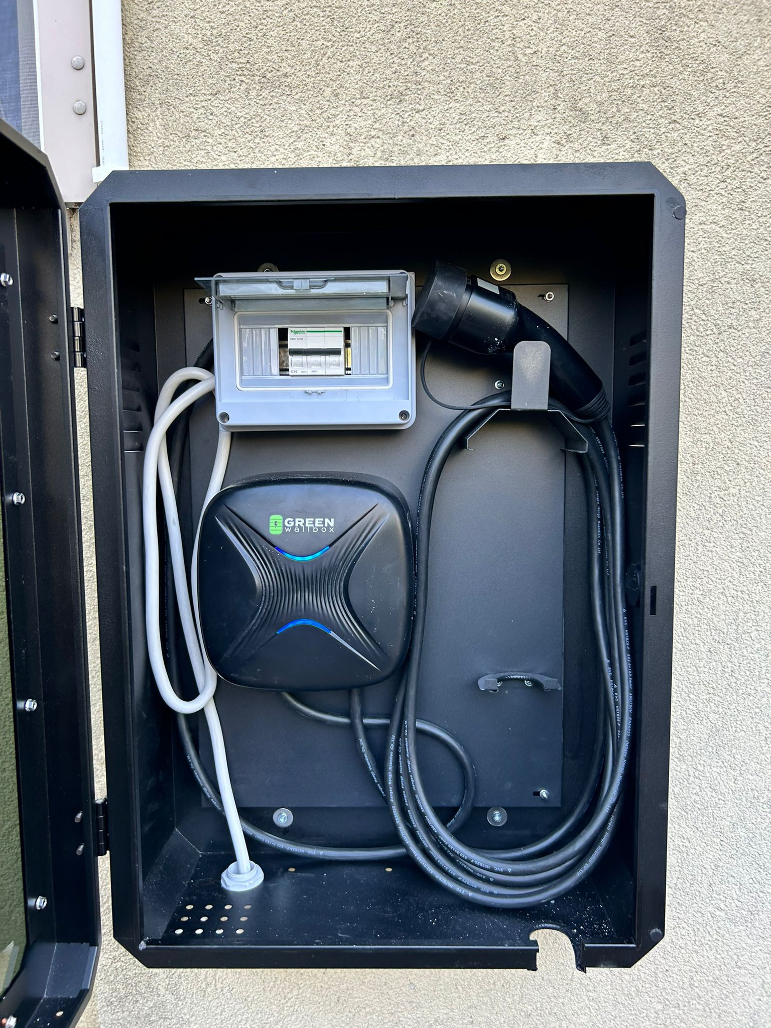 Czarna skrzynka z zamontowaną ładowarką do samochodu elektrycznego Green Wallbox i zabezpieczeniami elektrycznymi na szorstkiej ścianie zewnętrznej budynku.