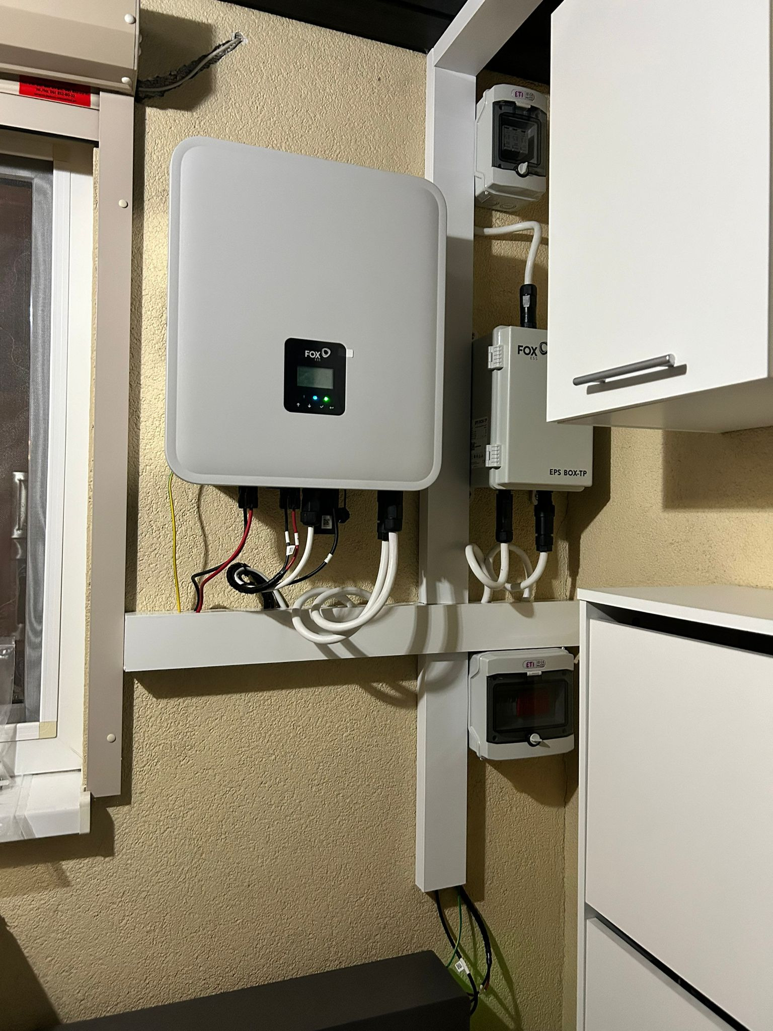 Wewnętrzny montaż falownika FOX ESS z podłączeniami kablowymi i zabezpieczeniami elektrycznymi na ścianie o chropowatej fakturze, obok okna i białych szafek.