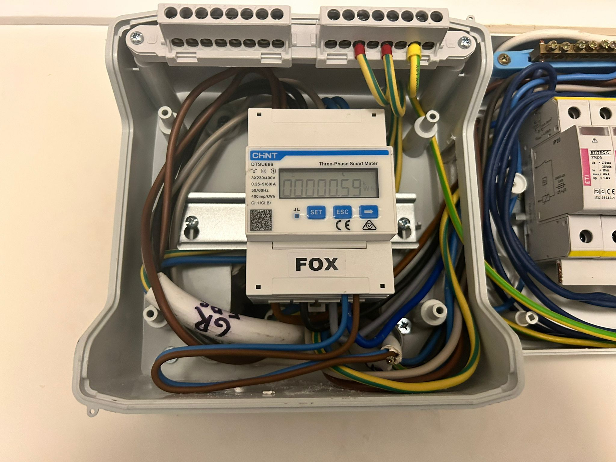 Otwarta skrzynka elektryczna z zamontowanym trójfazowym inteligentnym licznikiem energii elektrycznej firm CHNT i FOX, widoczne kable i zabezpieczenia.