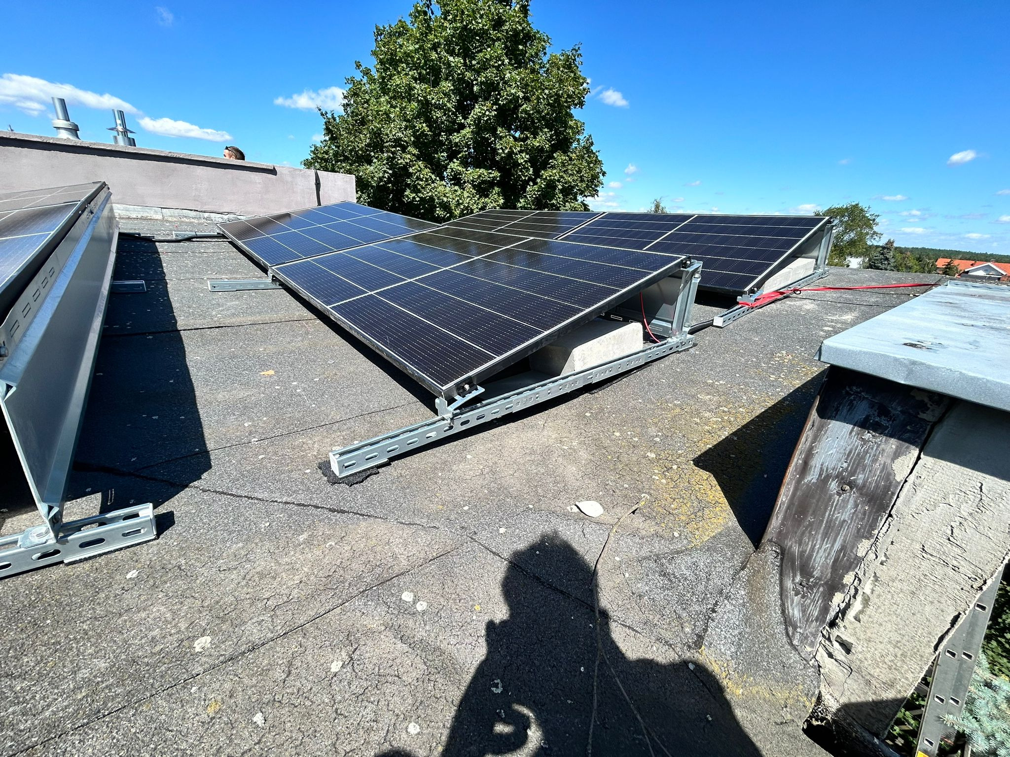 Panele słoneczne zamontowane na płaskim dachu budynku, widoczne szyny montażowe, betonowe podstawy i cień osoby robiącej zdjęcie.