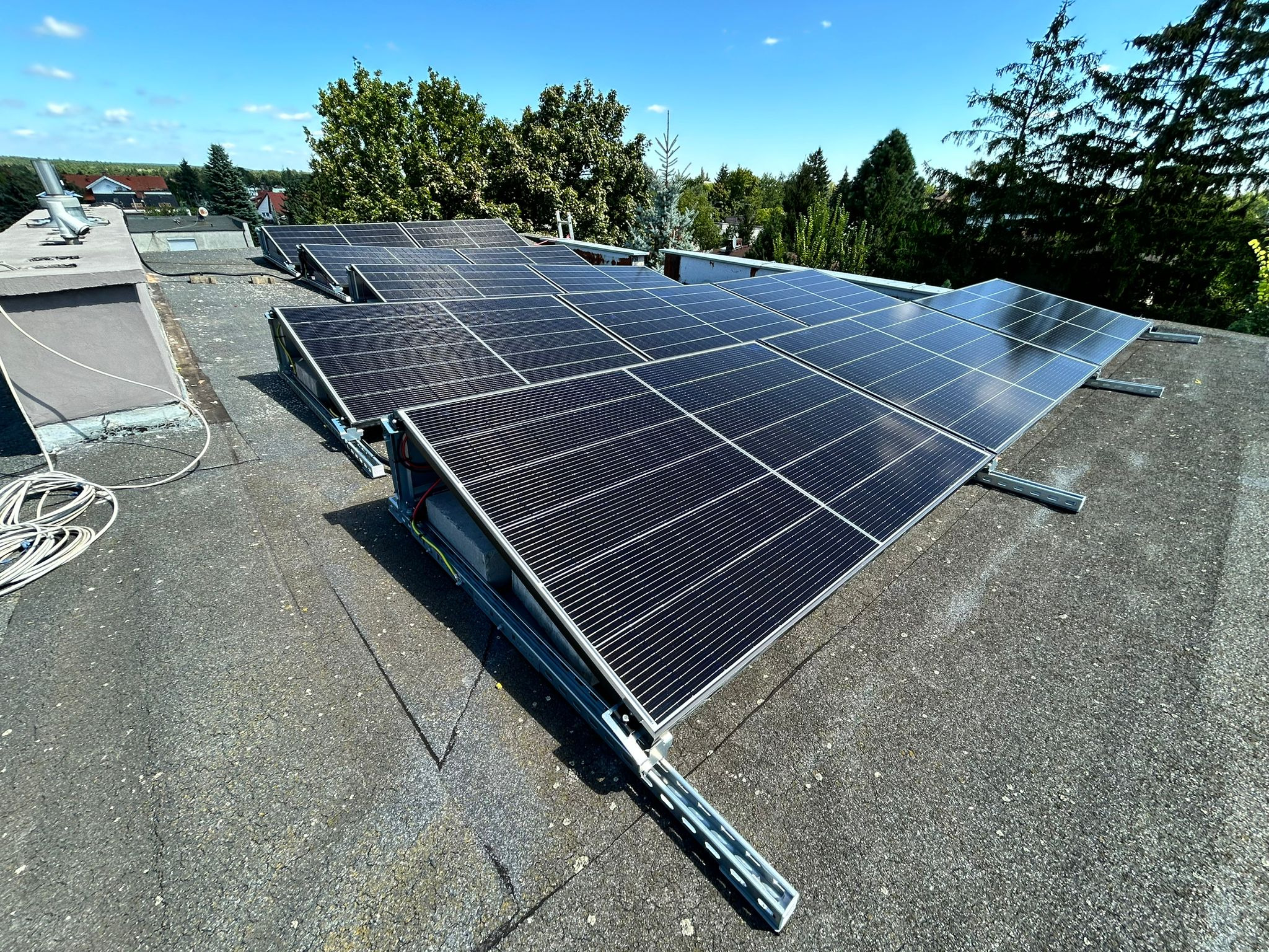 Rząd paneli fotowoltaicznych na płaskim dachu, mocowane na szarej konstrukcji wsporczej, z zielenią drzew w tle i niebieskim niebem.