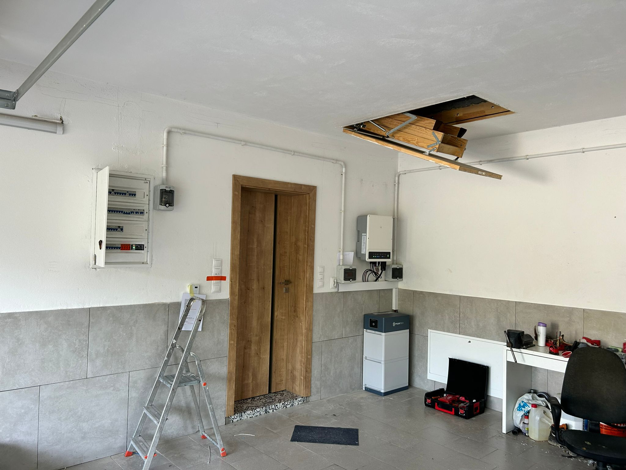 Wnętrze garażu z zainstalowanym akumulatorem energii, inwerterem i rozdzielnicą elektryczną; widoczna drabina, otwarta klapa strychowa i narzędzia na stole.