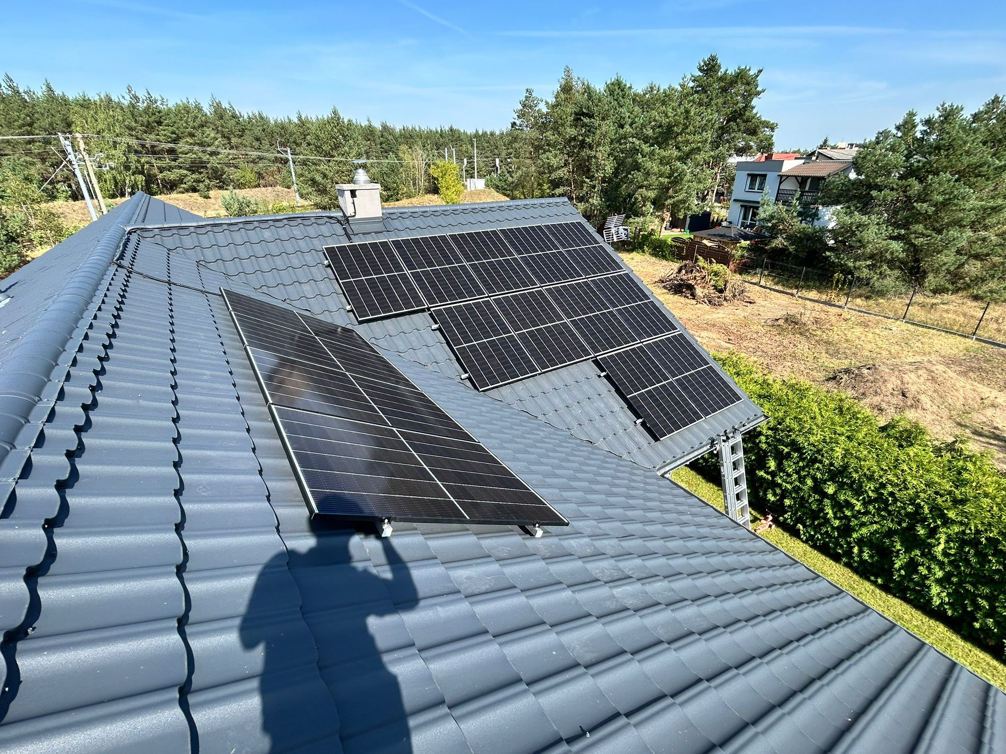Panele słoneczne na grafitowym dachu domu jednorodzinnego w Szczytnikach, cień fotografa widoczny na pierwszym planie.
