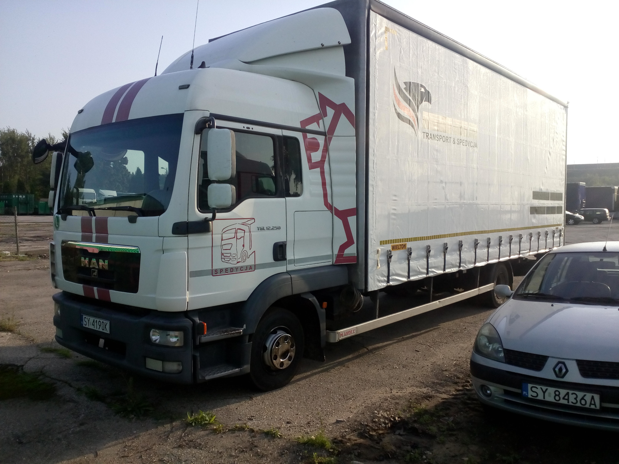 Ciężarówka MAN z naczepą z logo firmy transportowej, zaparkowana na placu, obok zaparkowany srebrny samochód osobowy.