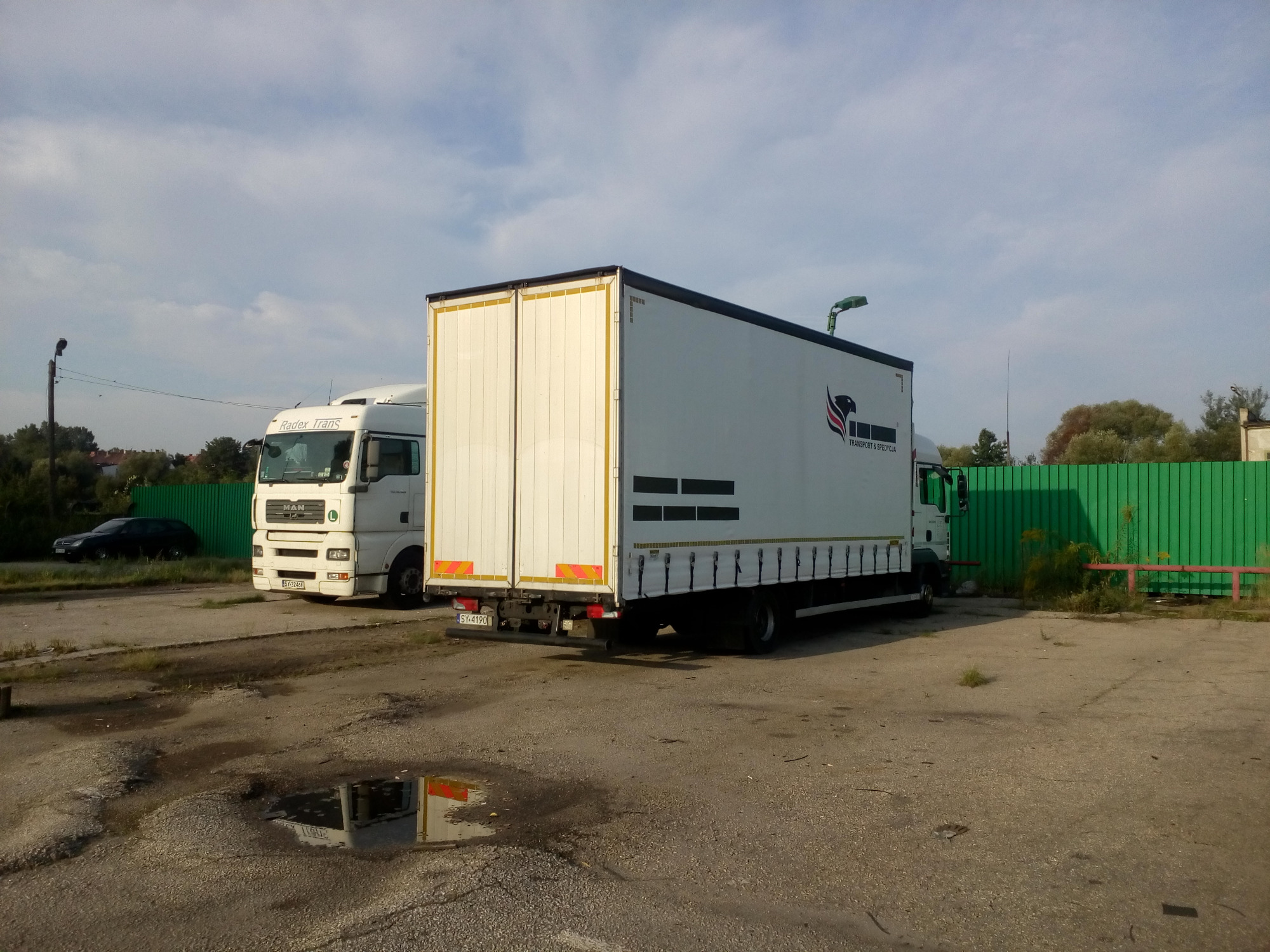 Dwa białe samochody ciężarowe zaparkowane na placu, jeden z widocznym logo firmy transportowej, drugi z widocznym napisem Radex Trans na kabinie, na tle zielonego ogrodzenia i pochmurnego nieba.