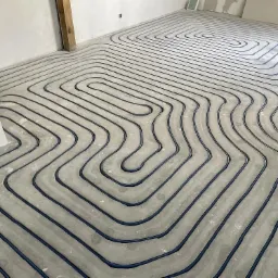 Układ ogrzewania podłogowego z niebieskich rur na szarym podłożu betonowym, widoczne niedokończone ściany i drewniany element konstrukcyjny