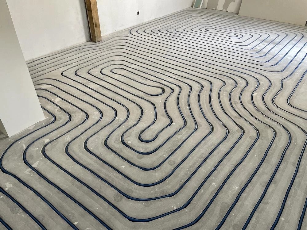 Układ ogrzewania podłogowego z niebieskich rur na szarym podłożu betonowym, widoczne niedokończone ściany i drewniany element konstrukcyjny