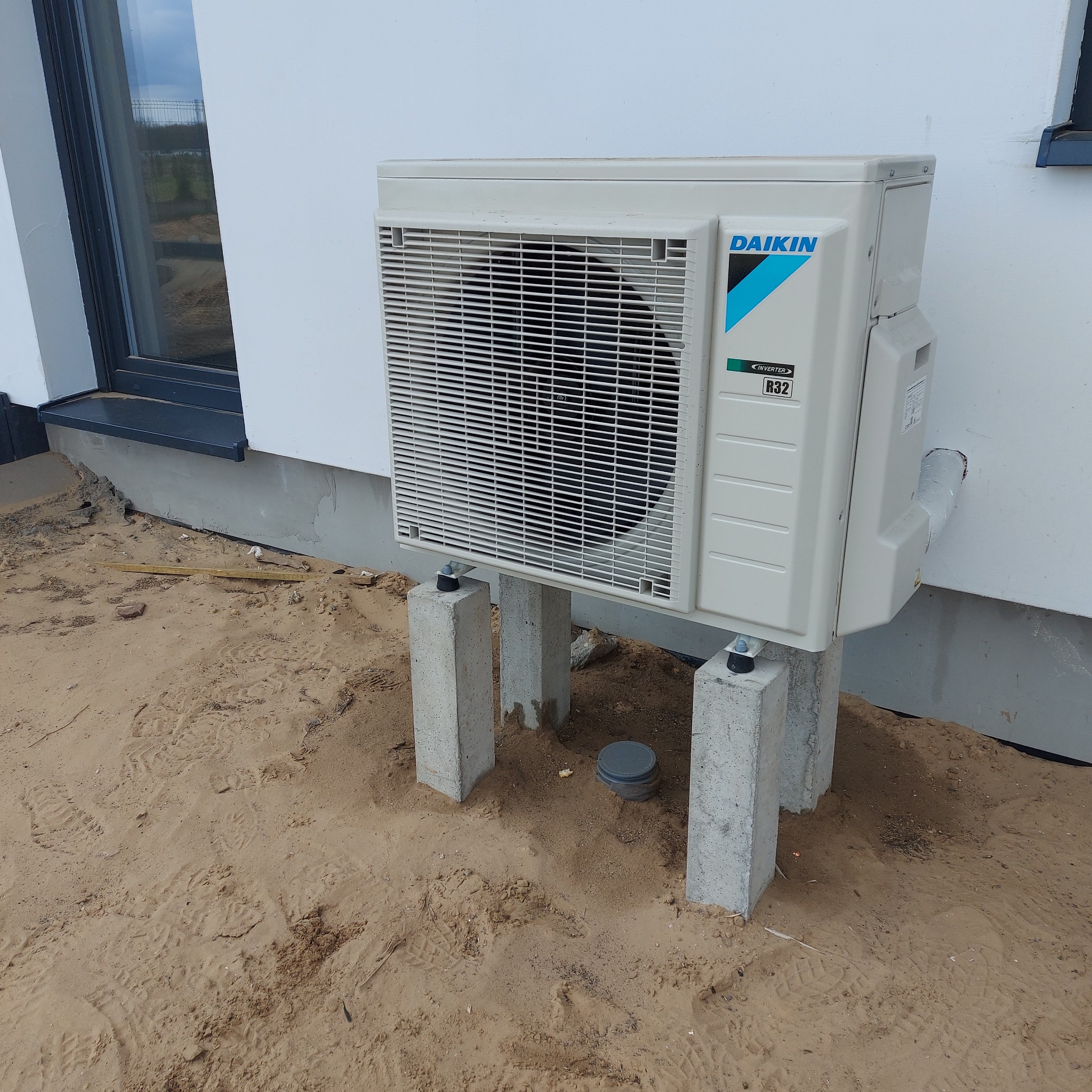Zewnętrzna jednostka klimatyzacji marki Daikin, model R32, zamontowana na betonowych wspornikach przy białej ścianie budynku, widoczne ślady prac ziemnych w postaci piasku.