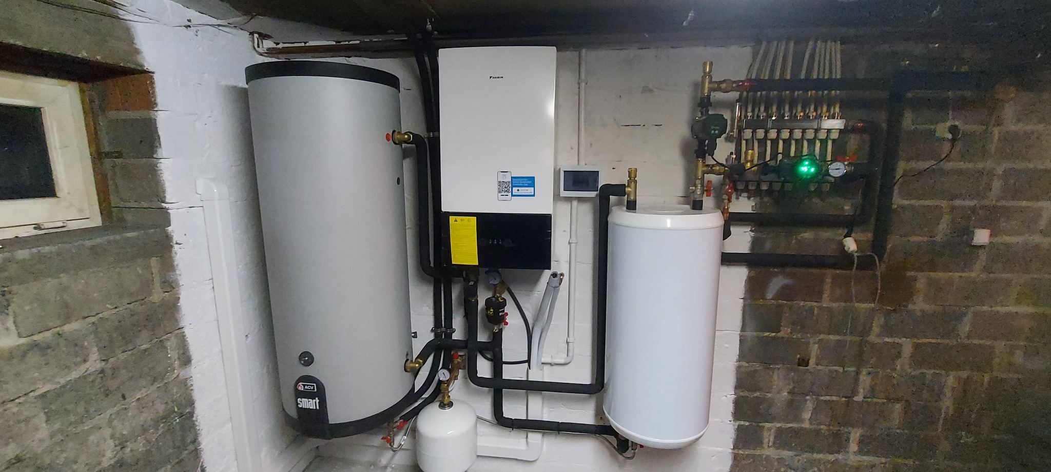 Zestaw urządzeń grzewczych w piwnicy z ceglanymi ścianami, zawierający biały zasobnik ciepłej wody użytkowej ACV Smart, jednostkę centralną Daikin z wyświetlaczem i rozdzielacz instalacji grzewczej...