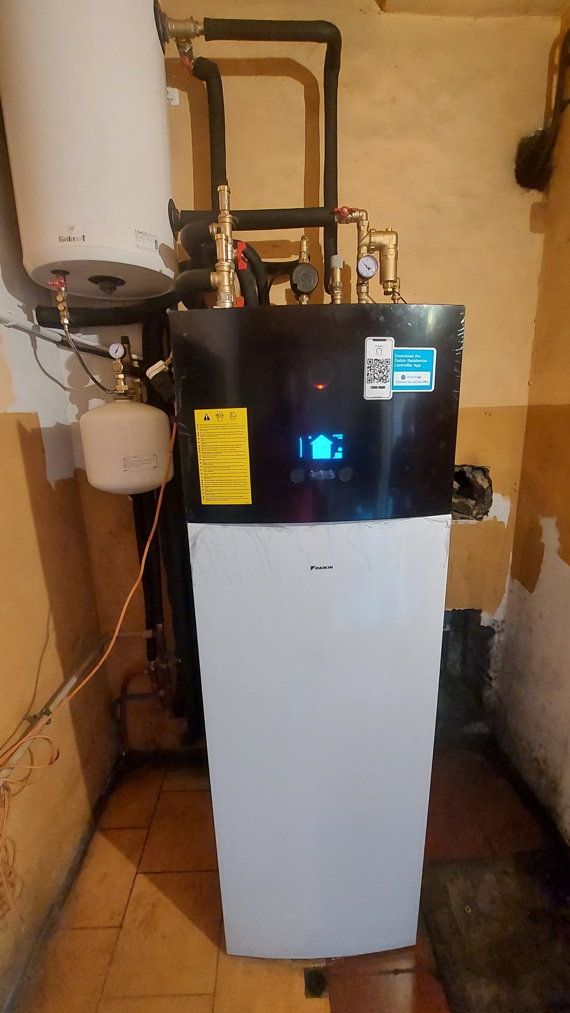Nowa, częściowo ofoliowana pompa ciepła Daikin, z podłączonymi rurami i zbiornikiem, zainstalowana w pomieszczeniu z pomarańczowymi ścianami i brudną podłogą.