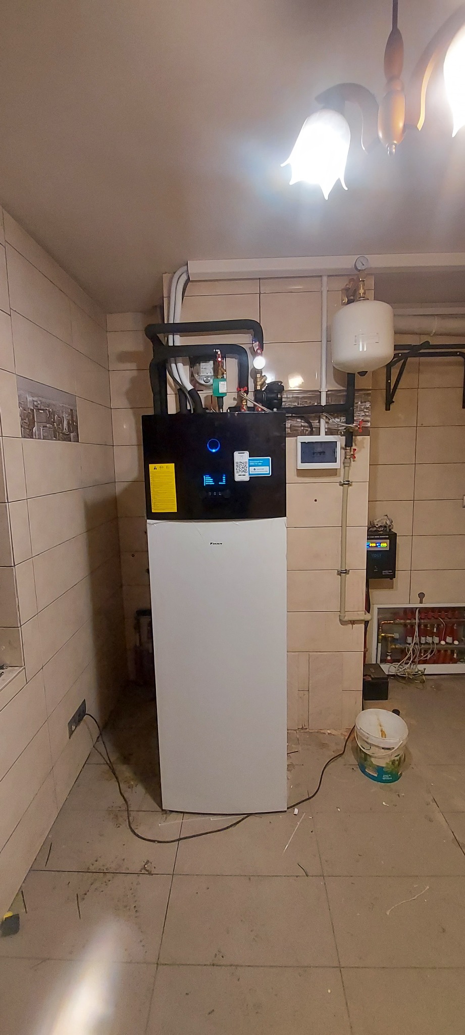Biała pompa ciepła Daikin z czarnym panelem sterowania, rury instalacyjne na ścianie z beżowych płytek, widoczny osprzęt i wiązka kabli, podłoga z zabrudzeniami po instalacji.